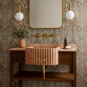 May include: A bathroom vanity with a fluted, reddish-pink marble sink. The sink sits on a dark wood cabinet with a matching countertop. A gold-framed mirror hangs above, with gold fixtures and a floral wallpaper background. Two white globe pendant lights hang above.