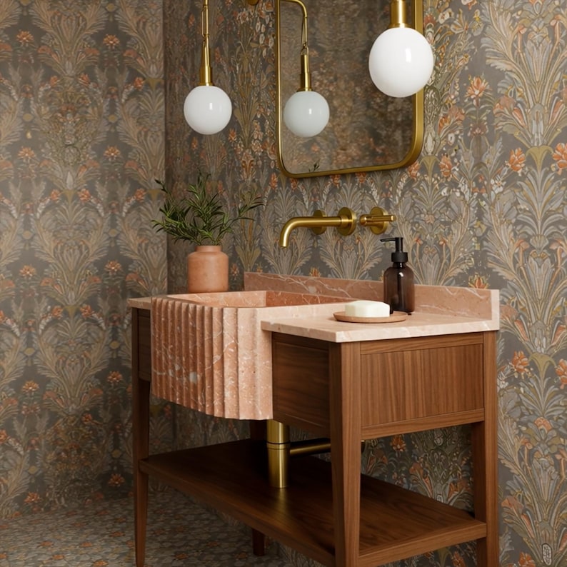May include: A bathroom vanity with a pink marble sink and a wooden cabinet. The faucet and mirror frame are gold. The wallpaper has a floral pattern in shades of brown, orange, and green. Two globe lights hang above the sink.