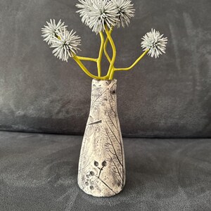 Könnte beinhalten: Eine kleine, cremefarbene Keramikvase mit einem Blättermuster und einem Strauß stacheliger, grauer Blumen mit gelben Stielen. Die Vase hat einen schmalen Hals und einen breiteren Boden. Die Blumen sind oben geclustert.