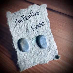 May include: Two smooth, oval-shaped, gray-blue stones rest on a piece of textured, light gray paper. The paper has the handwritten words "I'm Perfect Made" in black ink. The background is a wooden surface.