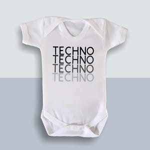 May include: White baby bodysuit featuring the word "TECHNO" printed in black, fading to grey. The short-sleeved onesie has a round neckline and snap closures at the bottom. The background is a two-tone grey and white.