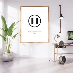 May include: A framed print with a black pause symbol and the text "Meet me in the present moment. Riss." The artwork is set in a room with a desk, a plant, and a fan. The frame is a light brown colour.