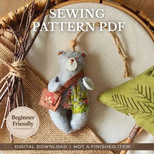 Può includere: Ornamento a forma di orso in feltro grigio con una borsa marrone, ricamata con fiori e foglie. Il testo "SEWING PATTERN PDF" è in alto. Il testo "Beginner Friendly" è in un cerchio in basso a sinistra. Il testo "DIGITAL DOWNLOAD | NOT A FINISHED ITEM" è in basso.
