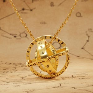Könnte beinhalten: Eine goldfarbene Armillarsphären-Anhängerhalskette mit mehreren ineinandergreifenden Ringen, die mit schwarzer Schrift und Tierkreiszeichen graviert sind. Der kugelförmige Anhänger hängt an einer feinen goldfarbenen Kette und zeigt ein komplexes, drehbares Design mit sichtbaren Schriftzeichen.