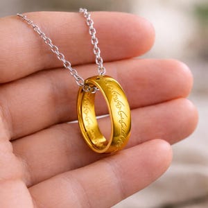 May include: A gold-colored ring pendant on a silver chain. The ring has engraved text and is held in a hand. The pendant is a circular band, and the chain is made of small, linked segments.