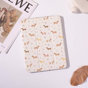 May include: A rectangular tablet case with a cream-colored background featuring a repeating pattern of small brown dachshund dogs in various poses, some wearing sweaters, interspersed with tiny colorful floral accents across the surface.