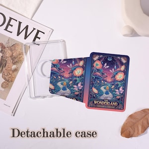May include: A clear, detachable phone case with a Wonderland-themed design. The design features a path, flowers, and the text "Visit Wonderland". The case is shown with a magazine and a decorative object.