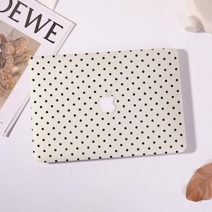 May include: A cream-coloured laptop case featuring a pattern of evenly spaced black polka dots. The Apple logo is visible in the centre. The case is on a white surface with a magazine and a decorative object in the background.