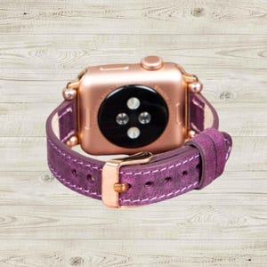 K&ouml;nnte beinhalten: Ros&eacute;goldene Apple Watch mit einem tiefvioletten Lederarmband. Das Armband hat wei&szlig;e N&auml;hte und eine goldene Schnalle. Die Uhr wird auf einer hellen Holzmaserung pr&auml;sentiert.