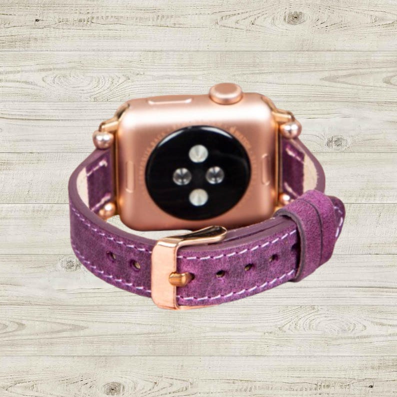 K&ouml;nnte beinhalten: Ros&eacute;goldene Apple Watch mit einem tiefvioletten Lederarmband. Das Armband hat wei&szlig;e N&auml;hte und eine goldene Schnalle. Die Uhr wird auf einer hellen Holzmaserung pr&auml;sentiert.
