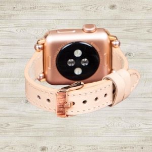 K&ouml;nnte beinhalten: Eine ros&eacute;goldene Smartwatch mit einem hellrosa Lederarmband. Das Uhrenarmband hat eine kupferfarbene Schnalle und wird mit einer Reihe von L&ouml;chern befestigt. Das Zifferblatt der Uhr ist schwarz.