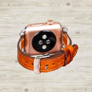 K&ouml;nnte beinhalten: Ros&eacute;goldene Smartwatch mit einem braunen Lederarmband. Das Armband ist mit wei&szlig;em Faden gen&auml;ht und hat eine silberne Schnalle. Die Uhr wird vor einem hellen Holzmaserungshintergrund gezeigt.
