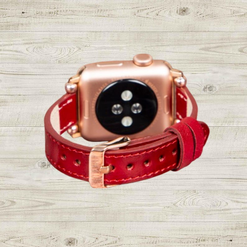 K&ouml;nnte beinhalten: Ros&eacute;goldene Apple Watch mit rotem Lederarmband. Das Armband hat eine goldfarbene Schnalle. Die Uhr wird auf einer hellen Holzmaserungsoberfl&auml;che pr&auml;sentiert.