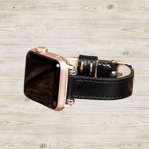 K&ouml;nnte beinhalten: Eine ros&eacute;goldene Apple Watch mit einem schwarzen Lederarmband. Das Zifferblatt der Uhr ist schwarz und das Armband hat wei&szlig;e N&auml;hte. Die Uhr wird auf einer hellen Holzmaserung pr&auml;sentiert.