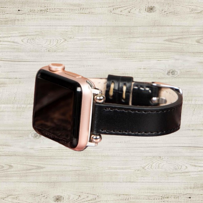 K&ouml;nnte beinhalten: Eine ros&eacute;goldene Apple Watch mit einem schwarzen Lederarmband. Das Zifferblatt der Uhr ist schwarz und das Armband hat wei&szlig;e N&auml;hte. Die Uhr wird auf einer hellen Holzmaserung pr&auml;sentiert.