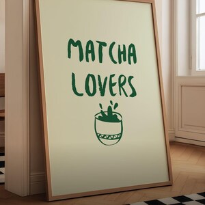 May include: A framed art print featuring the text "MATCHA LOVERS" in dark green brush lettering, with a green matcha bowl illustration below. The print is in a light-coloured frame and leans against a wall.