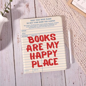 May include: A rectangular notebook with a cream-colored cover designed like a vintage library checkout card. It features blue horizontal lines, columns for author, title, and dates, and the bold red text: BOOKS ARE MY HAPPY PLACE.