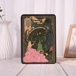 May include: A black decorative plaque featuring artwork from the musical "Wicked." The design includes a princess in a pink gown, a green witch, and a green castle. The word "Wicked" is prominently displayed in the center of the plaque.