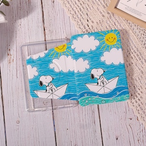 May include: A rectangular protective case for an electronic device featuring a blue sky background with white clouds and a yellow sun. The design depicts a white cartoon dog with black ears sitting in a white paper boat on blue water.