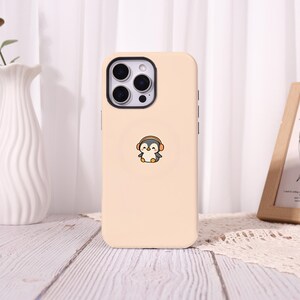 May include: A light beige smartphone case featuring a small, centred illustration of a cartoon penguin wearing orange headphones. The case has a black camera module cutout with three circular lenses and a circular MagSafe-style ring outline.