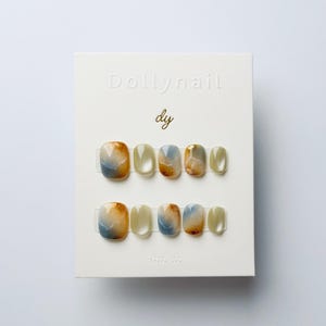 Könnte beinhalten: Ein Set künstlicher Fingernägel auf einer weißen Karte. Die Nägel haben eine quadratische Form mit einem marmorierten Design in Blau, Gold und Beige. Auf der Karte sind die Texte "Dollynail" und "dy" geprägt.