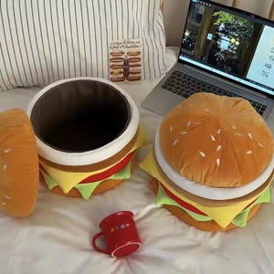Puede incluir: Una almohada y un contenedor de almacenamiento con forma de hamburguesa de felpa con diseño de pan, carne, queso y lechuga. También hay una taza roja con pequeñas ilustraciones y un ordenador portátil. La almohada de hamburguesa mide unos 30 cm de diámetro.