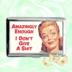 Puede incluir: Un estuche de metal plateado con el texto "AMAZINGLY ENOUGH I DON'T GIVE A SHIT" en rojo. El estuche presenta una ilustración vintage de una mujer con ojos azules y un peinado retro, junto con varias mariposas amarillas.