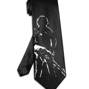 May include: Black necktie featuring a white silhouette of a person holding a guitar. The tie is made of a smooth, dark fabric and is suitable for formal or casual wear. The design is a bold, graphic statement.