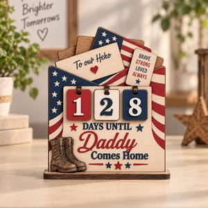 May include: A wooden countdown sign featuring a red, white, and blue American flag design. It displays the text To our Heko, BRAVE STRONG LOVED ALWAYS, and 128 DAYS UNTIL Daddy Comes Home. The sign includes three rotating number tiles and a pair of brown military-style boots.