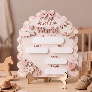 Può includere: Una targa nascita in legno smerlato con il testo "hello World" e "my name is :", dotata di spazi vuoti per data, ora, peso e lunghezza. La targa è decorata con un fiocco rosa, nuvole, stelle, fiori e cuori in tonalità delicate.