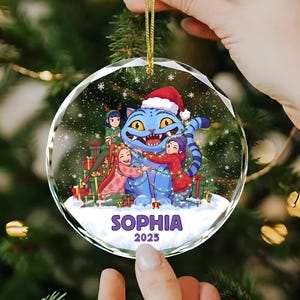 May include: A clear, round Christmas ornament featuring a blue cat wearing a Santa hat, surrounded by three people hugging it. The ornament has the name "SOPHIA" and the year "2025" printed on it. The ornament is hanging from a gold string.
