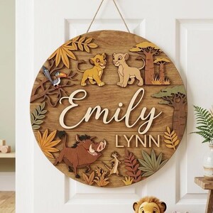 May include: A round wooden sign featuring a Lion King theme. The sign includes the name "Emily Lynn" in a cursive font, along with illustrations of Simba, Nala, Timon, Pumbaa, and Zazu. The sign is hung with a jute rope.