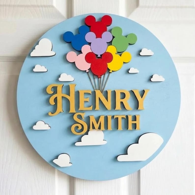 May include: A round, light blue wooden sign with the name "HENRY SMITH" in gold lettering. Colorful Mickey Mouse-shaped balloons are attached to the top, with white clouds scattered around the sign.