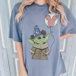 Puede incluir: Camiseta azul vaquero con un gr&aacute;fico de un personaje alien&iacute;gena verde que lleva un sombrero de mago con orejas de Mickey Mouse. El alien&iacute;gena sostiene un globo de Mickey Mouse rosa. El texto "BLUE JEAN" est&aacute; en la parte inferior.
