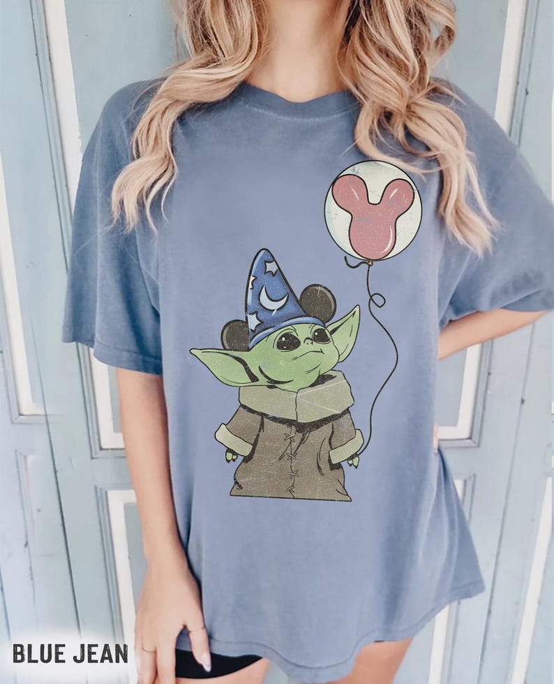 Puede incluir: Camiseta azul vaquero con un gr&aacute;fico de un personaje alien&iacute;gena verde que lleva un sombrero de mago con orejas de Mickey Mouse. El alien&iacute;gena sostiene un globo de Mickey Mouse rosa. El texto "BLUE JEAN" est&aacute; en la parte inferior.