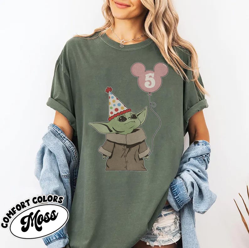 May include: Olive green t-shirt featuring a cartoon character wearing a party hat and holding a pink balloon with the number 5. The shirt has the words "COMFORT COLORS" and "Moss" printed on it.