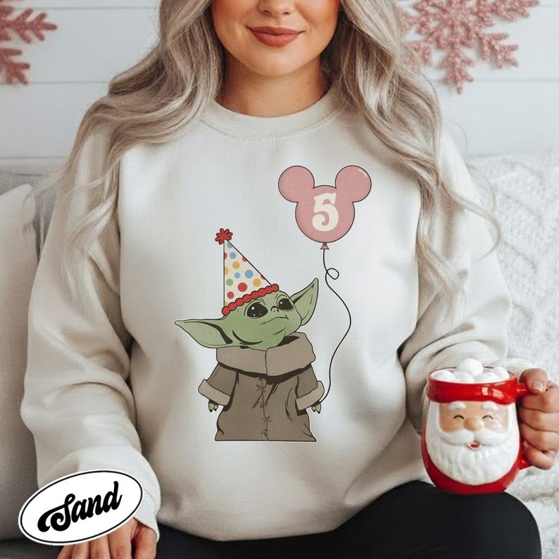 May include: A cream-colored sweatshirt featuring a cartoon illustration of a green alien character wearing a party hat and holding a pink balloon with the number 5. A red Santa mug is held in the hand.