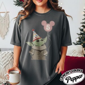 May include: Dark gray t-shirt featuring a cartoon character wearing a party hat and holding a pink balloon with the number 5. The shirt has the Comfort Colors Pepper logo.