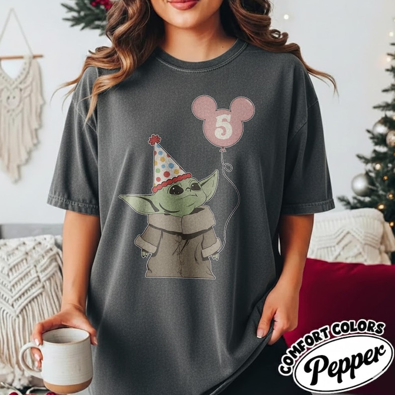 May include: Dark gray t-shirt featuring a cartoon character wearing a party hat and holding a pink balloon with the number 5. The shirt has the Comfort Colors Pepper logo.