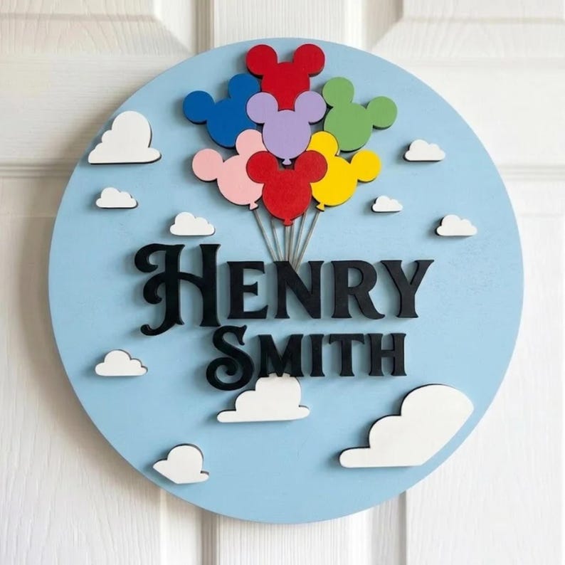 May include: A round, light blue wooden sign with the name "HENRY SMITH" in black letters. Above the name are colorful Mickey Mouse-shaped balloons. White cloud cutouts are scattered around the sign, creating a whimsical, decorative piece.