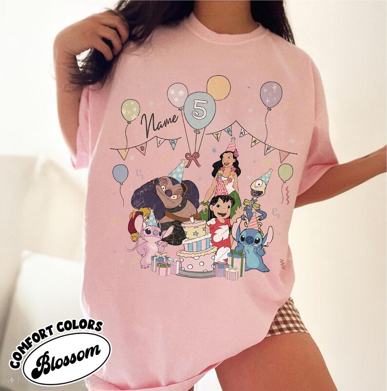 May include: Pink t-shirt featuring a colorful graphic of cartoon characters celebrating a birthday. The design includes balloons, a cake, and the number 5. The word "Name" is written above the characters.