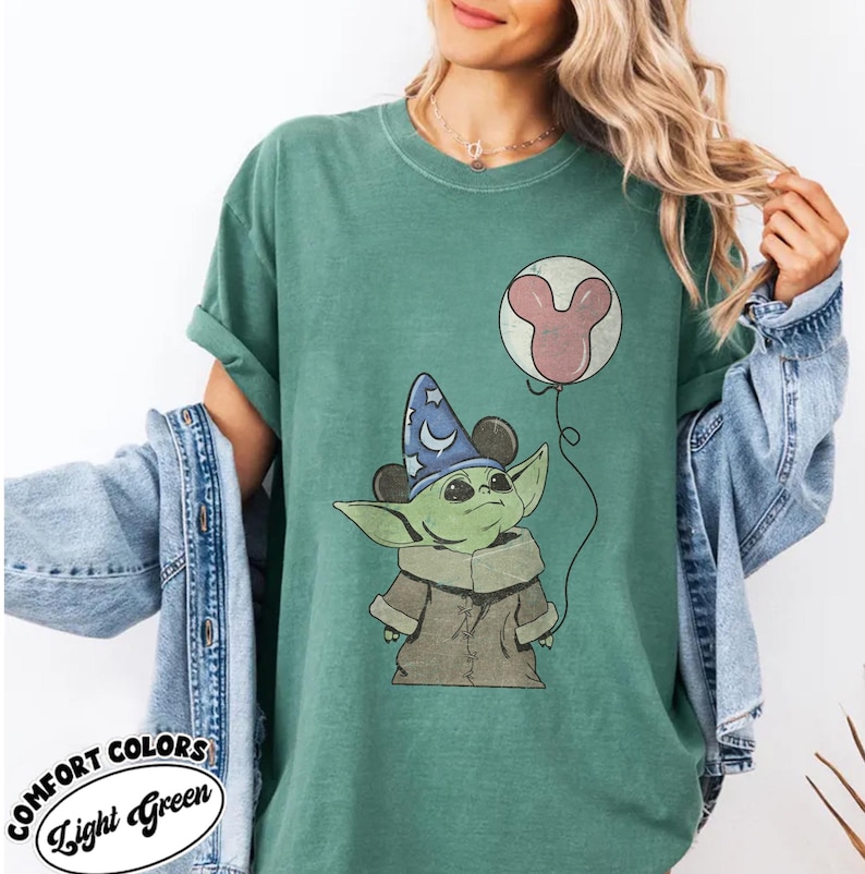 Puede incluir: Camiseta verde claro con un personaje de dibujos animados que lleva un sombrero de mago y sostiene un globo de Mickey Mouse. La camiseta tiene las palabras "COMFORT COLORS" y "Light Green".