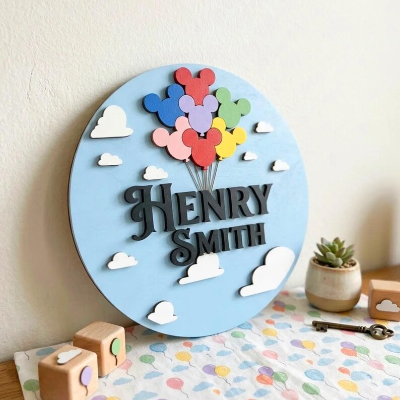 May include: A round, light blue wooden sign with the name "Henry Smith" in black, stylized lettering. Colorful Mickey Mouse-shaped balloons float above the name, and white clouds are scattered around the sign. A small succulent plant and wooden blocks are in the background.