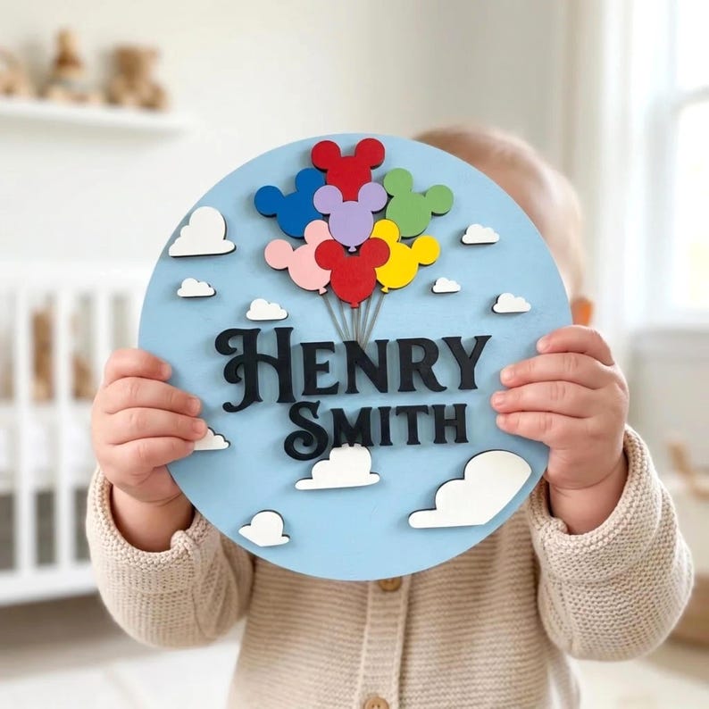 May include: A round, light blue wooden sign with the name "Henry Smith" in black script. The sign features colorful Mickey Mouse-shaped balloons and white cloud cutouts. A charming nursery or children's room decoration.