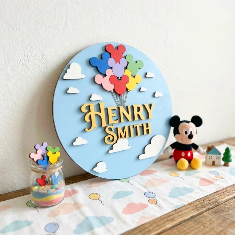 May include: A light blue wooden sign with the name "Henry Smith" in gold lettering, adorned with Mickey Mouse-shaped balloons in various colors and white cloud cutouts. A Mickey Mouse plush toy and a jar with Mickey Mouse cutouts are also present.