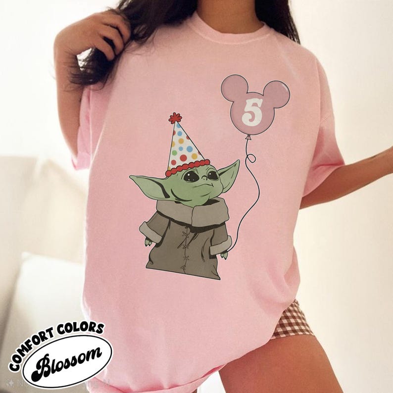 May include: Pink t-shirt featuring a cartoon illustration of a green alien character wearing a party hat and a gray coat. A pink Mickey Mouse-shaped balloon with the number 5 floats above the character. The text "COMFORT COLORS Blossom" is printed on the bottom left.