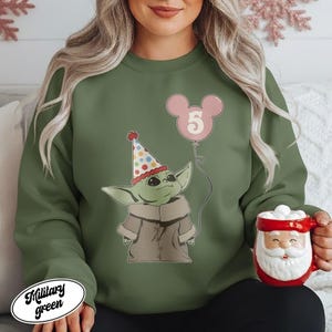 May include: Military green sweatshirt featuring a cartoon character wearing a party hat and holding a pink balloon with the number 5. The sweatshirt has a white oval label with the words "Military green". A Santa mug is held in the hand.