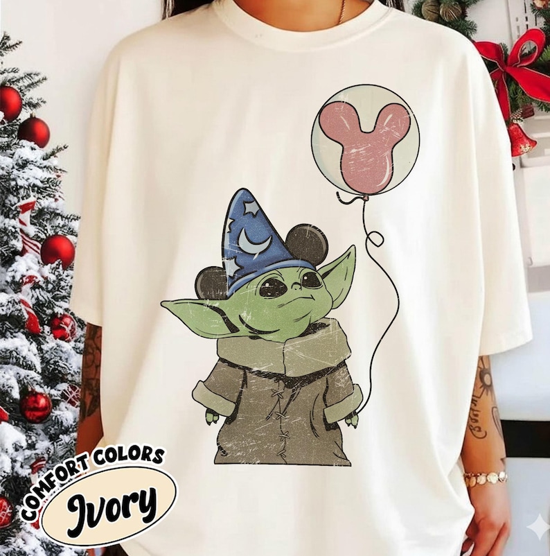 Puede incluir: Camiseta de color marfil con un personaje verde alien&iacute;gena que lleva un sombrero de mago azul con orejas de Mickey Mouse. El personaje sostiene un globo de Mickey Mouse rosa. La camiseta tiene las palabras "COMFORT COLORS" e "Ivory" impresas.