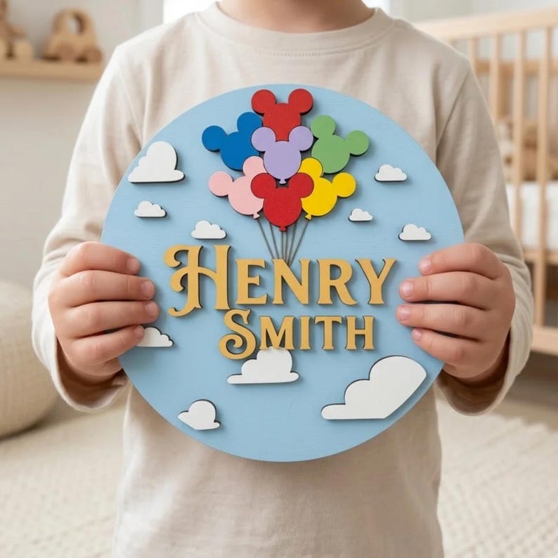 May include: A round, light blue wooden sign with the name "Henry Smith" in gold lettering. The sign features colorful balloon-shaped designs and white cloud cutouts. The sign is held by a person.