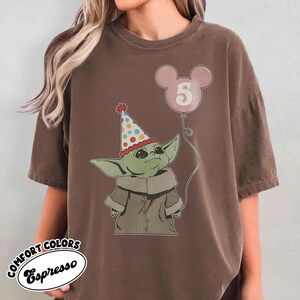 May include: Brown t-shirt featuring a graphic of a green alien-like character wearing a party hat and holding a pink balloon with the number 5. The shirt has the words "COMFORT COLORS Espresso" printed on it.